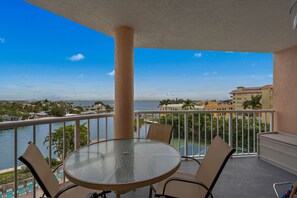 A balcony with a glass table and chairs, overlooking a body of water and a cityscape.