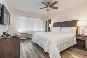 Quarto de hotel com uma cama grande, duas luminárias de cabeceira, um ventilador de teto, uma escrivaninha com telefone e uma janela com persianas.