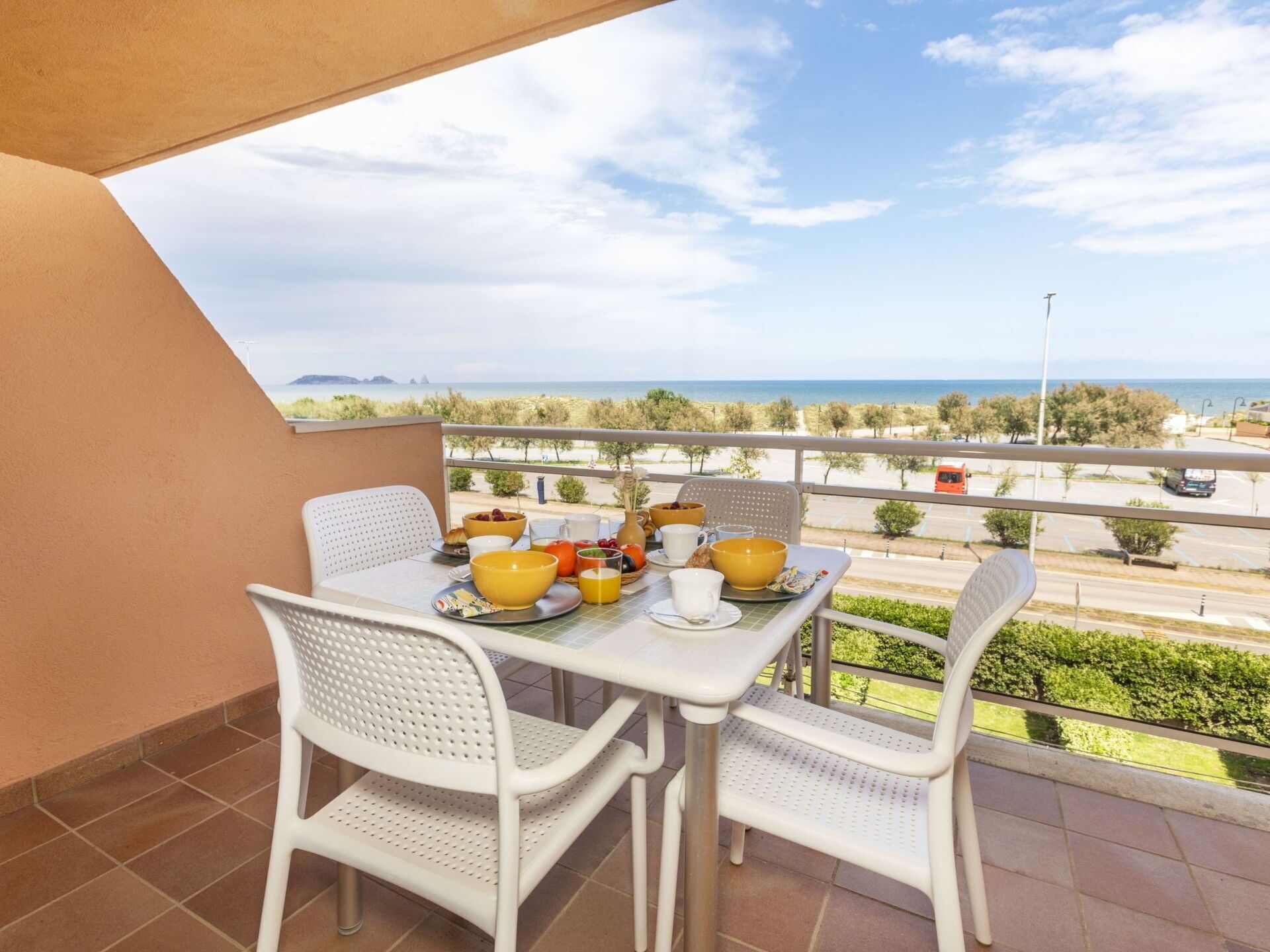 A balcony with a table set for breakfast, overlooking a beach and road.