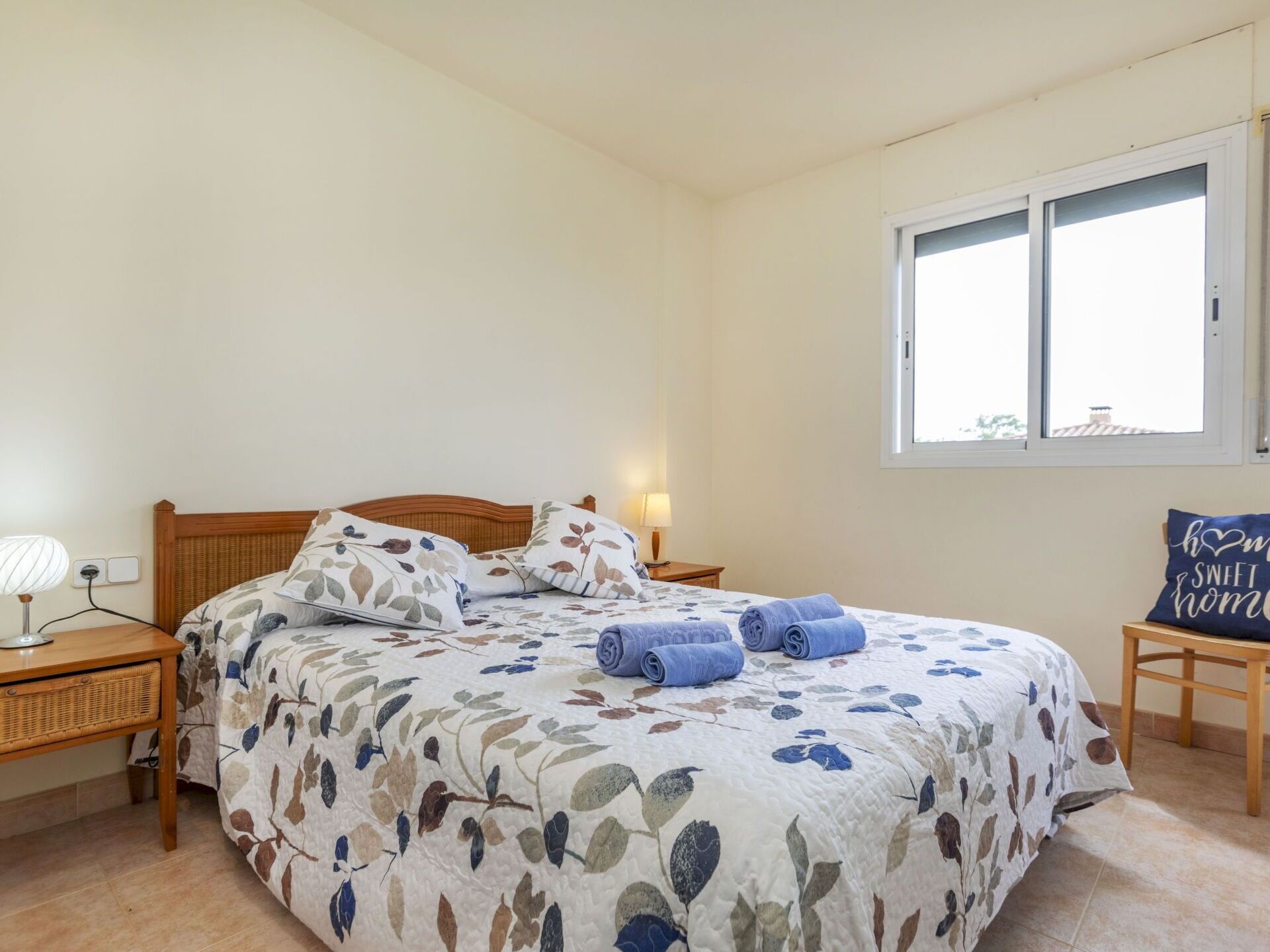 A bedroom with a floral bedspread, a wooden headboard, a bedside table with a lamp, and a window with a view.