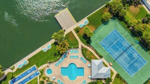 Vista aérea de un complejo turístico con una piscina central, múltiples tumbonas, cancha de tenis y muelle para embarcaciones.