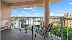Un balcon avec une table ronde et des chaises, donnant sur une étendue d’eau et des bâtiments.