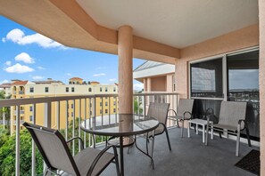 A balcony with a round glass table and four chairs, overlooking a cityscape with yellow buildings. - Elevated Penthouse | Bay Views & Perks (St. Pete Beach)