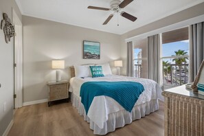 A bedroom with a bed, two bedside lamps, a ceiling fan, a window with curtains, and a view of the outside. - Elevated Penthouse | Bay Views & Perks (St. Pete Beach)