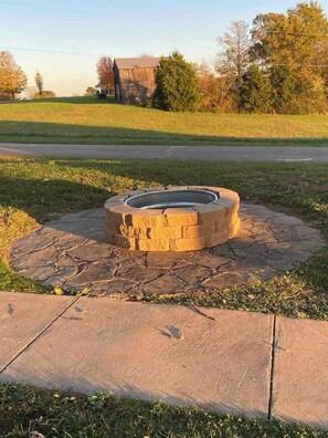 A circular stone fire pit with a metal rim, surrounded by a stone border, set in a grassy area with a paved path leading to it.