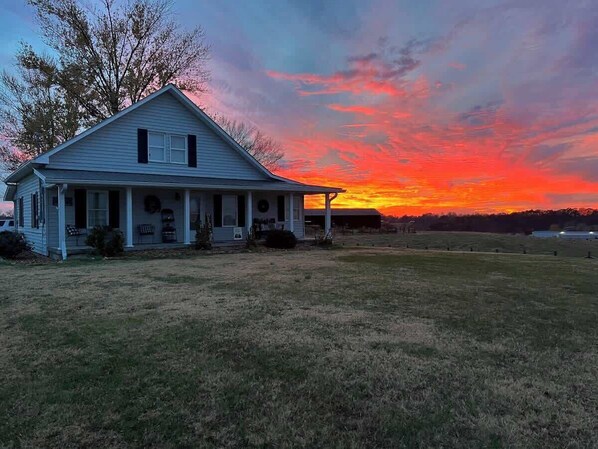 A single-story house with a porch, a front yard, and a sunset in the background.