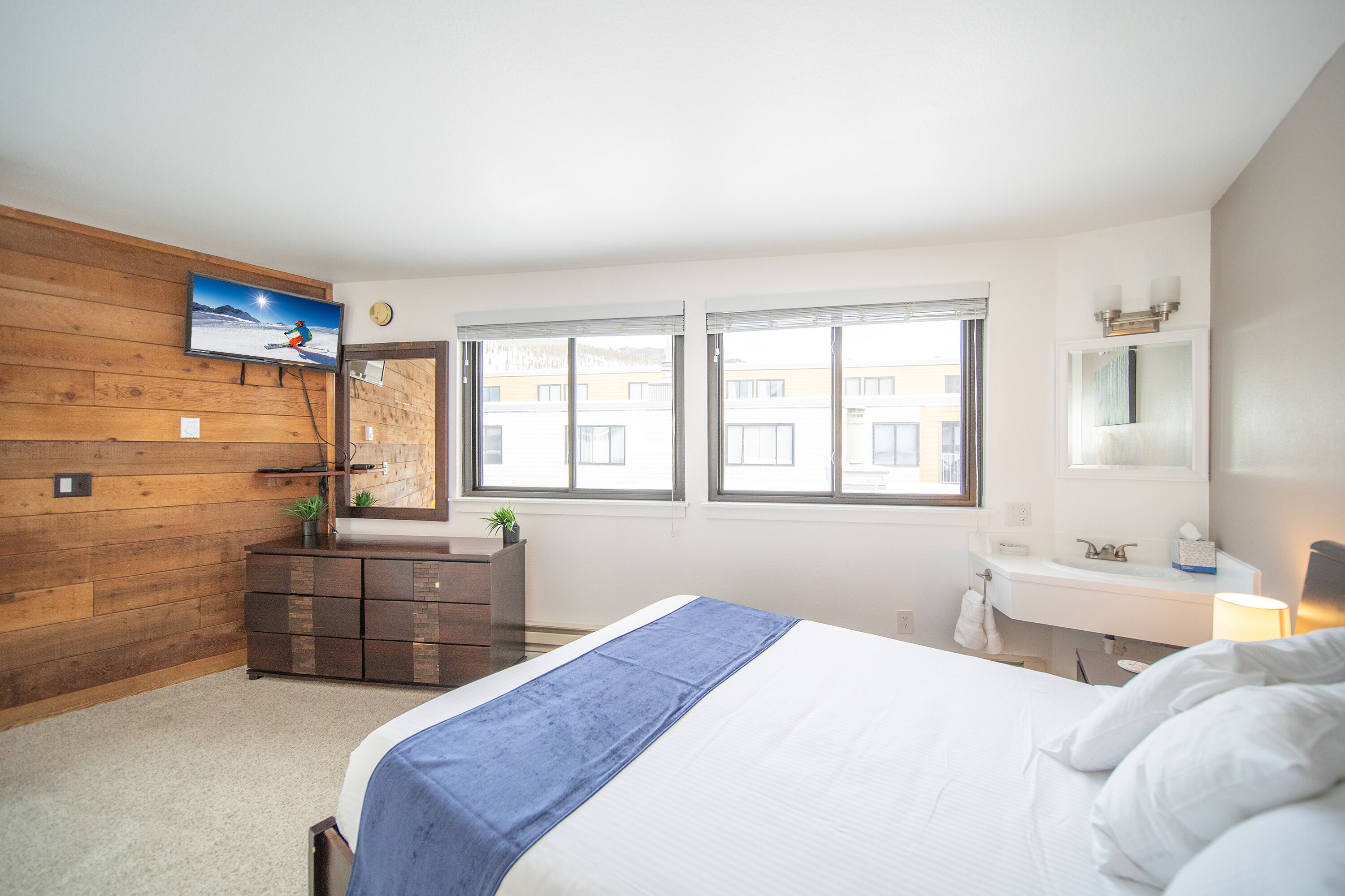 Un dormitorio con cama, cómoda, televisión montada en una pared de madera, ventana y baño con lavamanos y espejo.