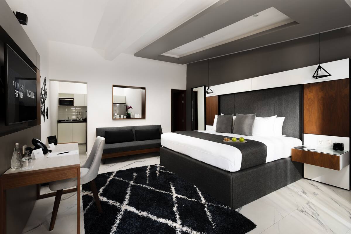 A modern bedroom with a large bed, a desk, a sofa, and a kitchen area in the background.