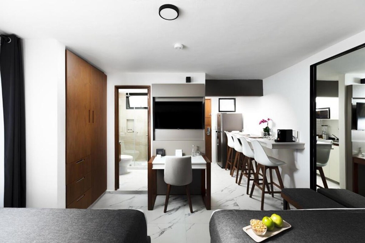 A modern hotel room with a dining area, a TV, and a kitchen.