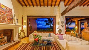 A living room with a fireplace, a sofa, a coffee table with flowers, and a view of the ocean at dusk.