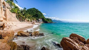 A beach with clear turquoise water, rocky shore, and a building with a stone facade.