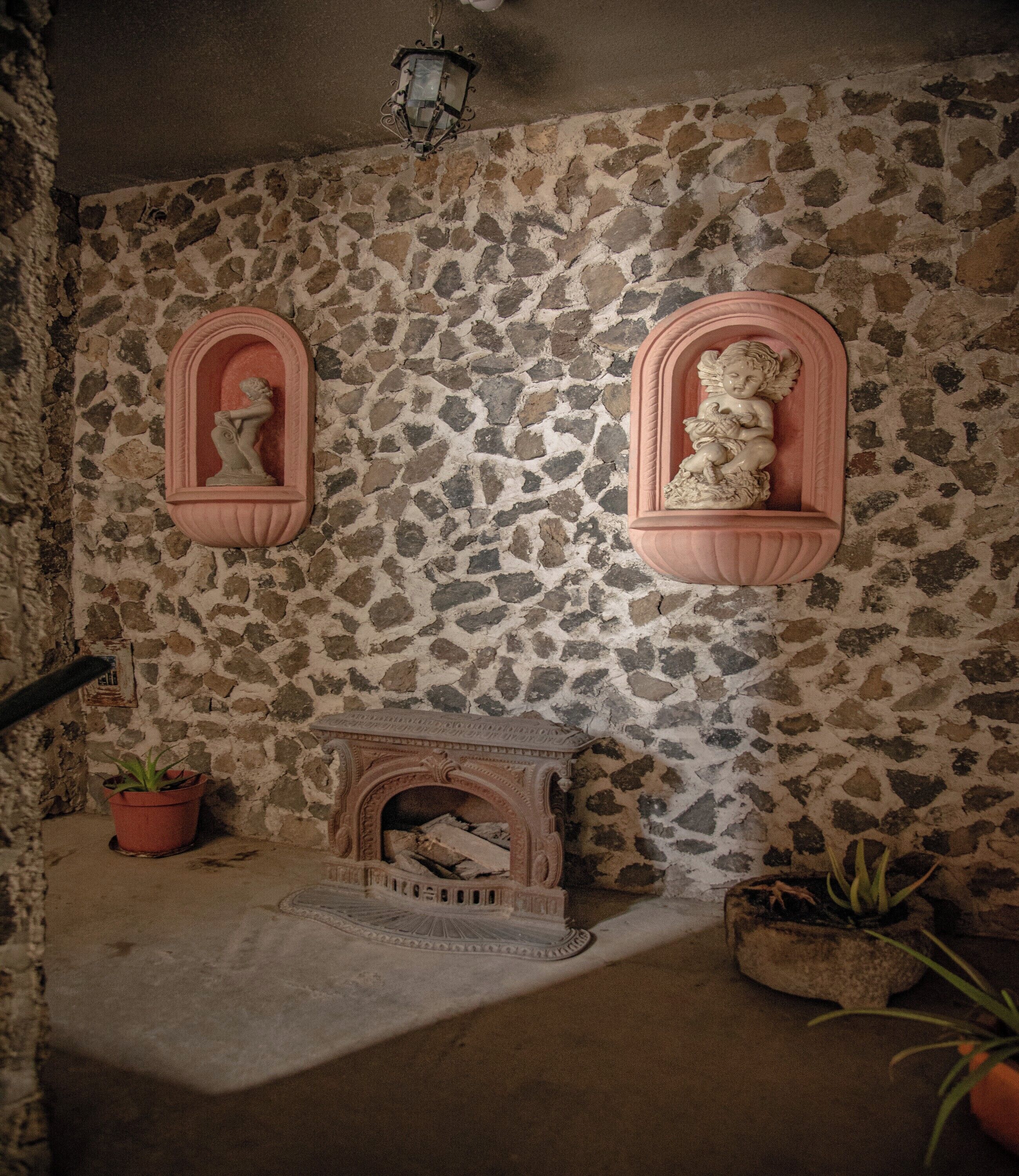 A stone wall with two pink-framed niches containing statues, a fireplace with a stone mantle, and potted plants.