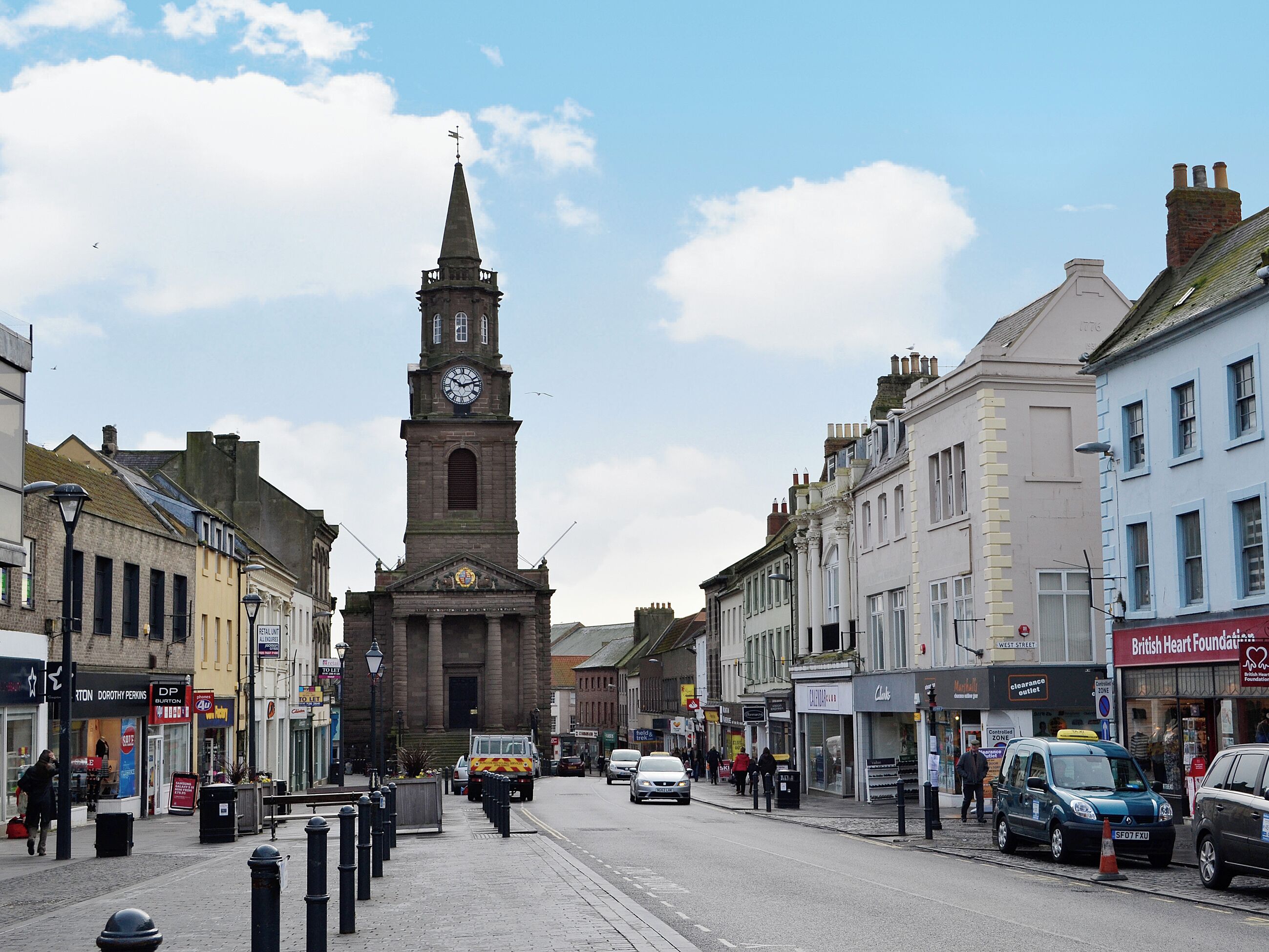 A street with a prominent clock tower, shops, and parked cars.