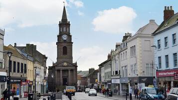 A street with a prominent clock tower, shops, and parked cars.