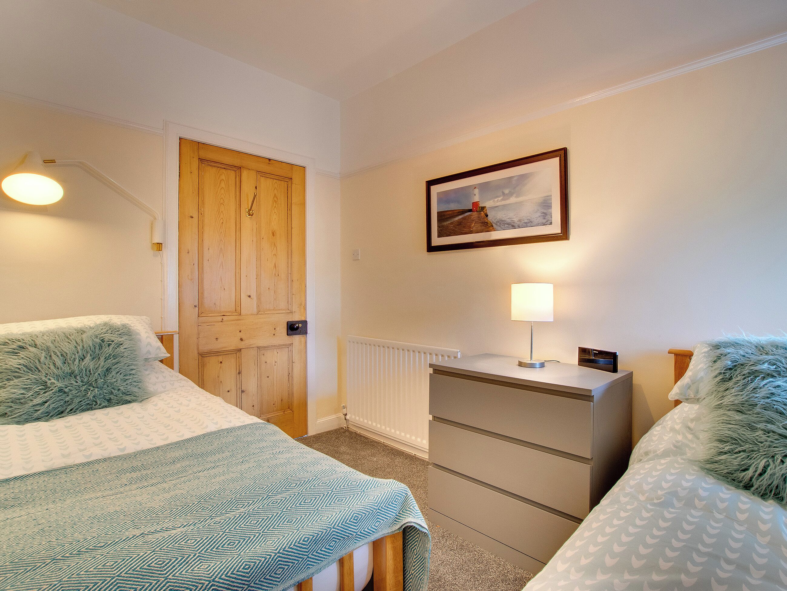 A bedroom with two beds, a dresser, a lamp, a framed picture on the wall, and a wooden door.
