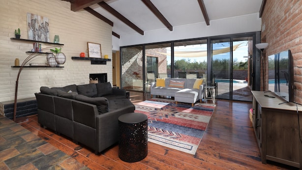 A spacious living area with a dark sofa, a patterned rug, and a wooden coffee table. There is a brick wall and a large sliding glass door leading to an outdoor patio. - Luxe & Spacious Gem: Pool ~ 15min to Downtown ~ Parking! (Tucson)