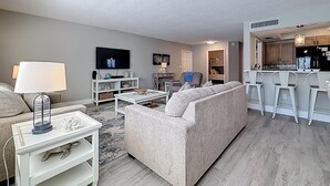 A modern living room with a sofa, coffee table, and TV. There is a kitchen area with bar stools and a refrigerator. - Poolside Patio | Walk to Beach (St. Pete Beach)