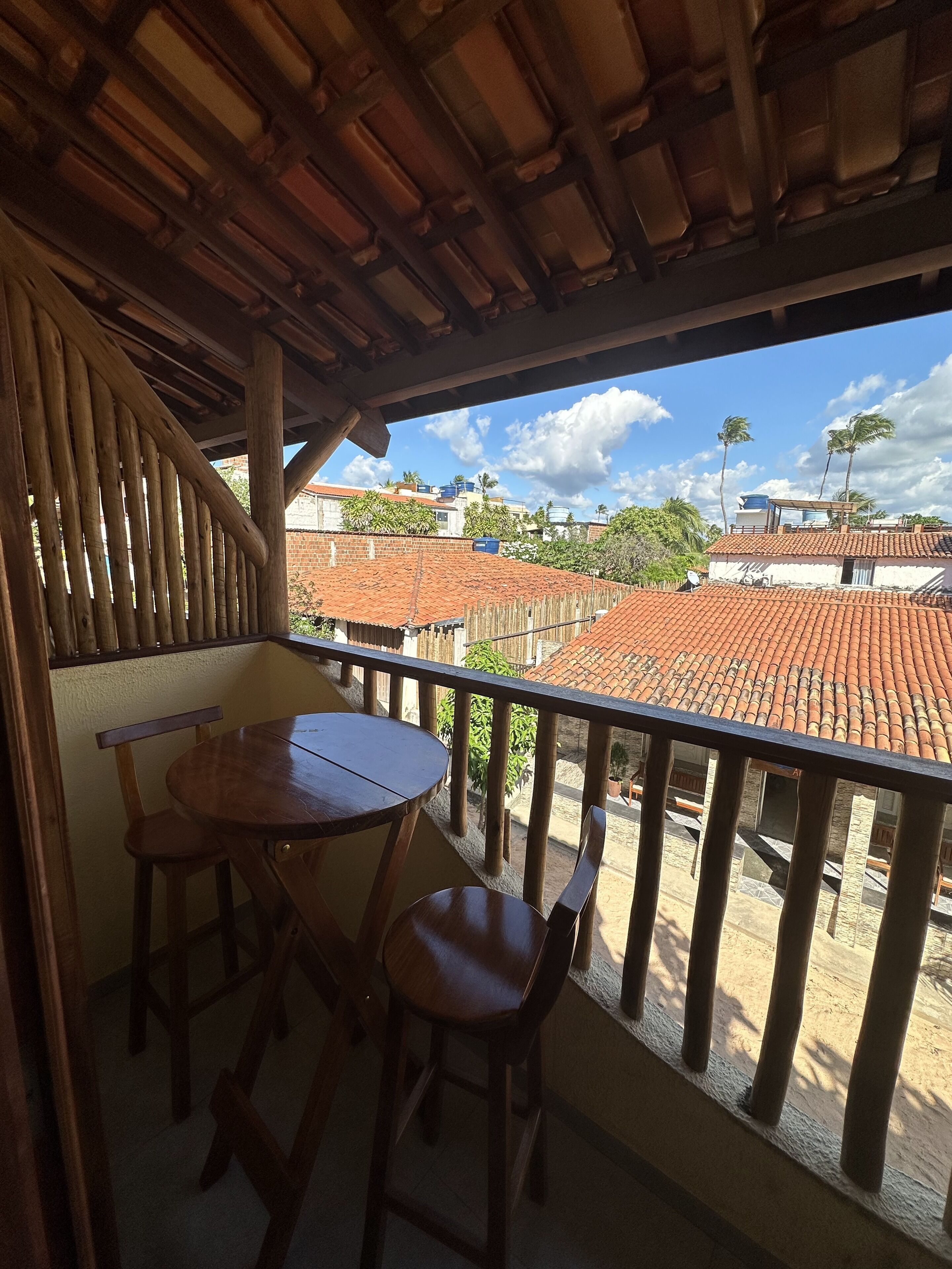 A balcony with a wooden table and chairs, overlooking a residential area with red-tiled roofs and palm trees.