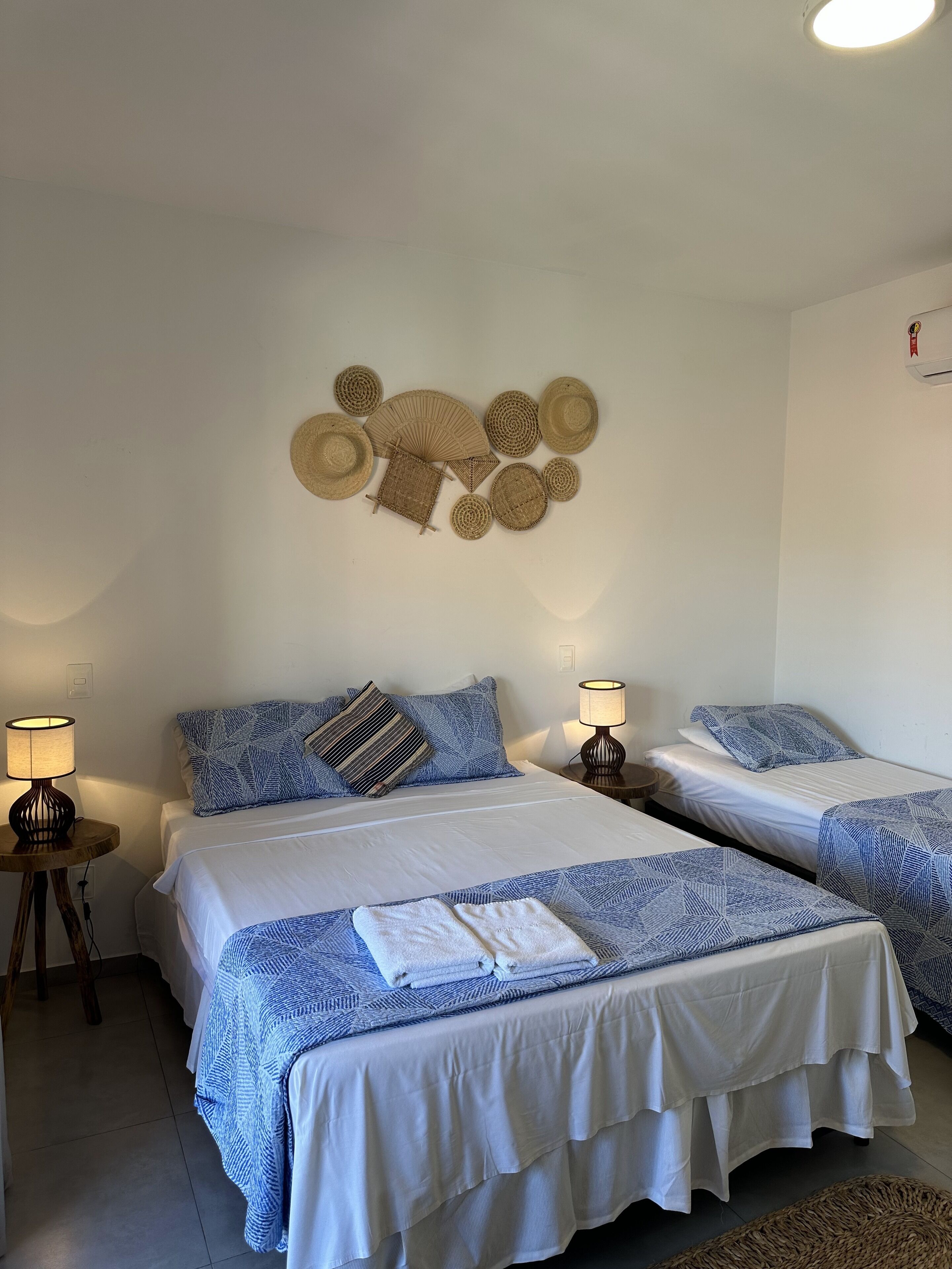 Quarto de hotel com duas camas, cada uma com colcha estampada em azul e branco, almofadas brancas e uma pequena mesa de cabeceira com abajur.
