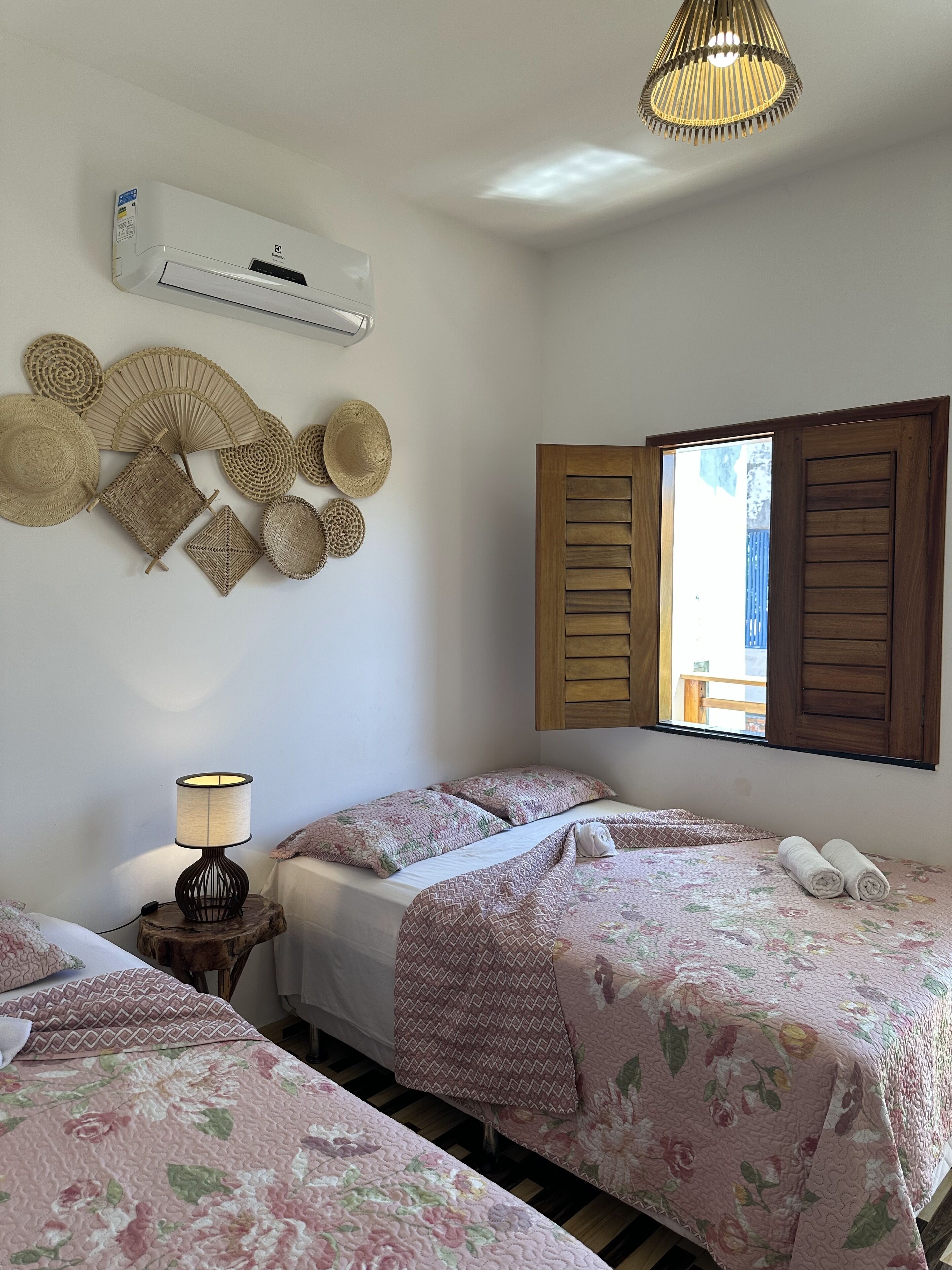 A bedroom with two beds, a wall-mounted air conditioner, a lamp, and a window with wooden shutters.