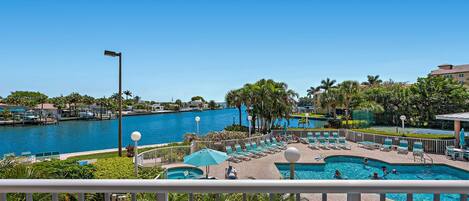 A pool area with lounge chairs and umbrellas, surrounded by palm trees and a clear blue sky.
