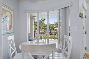 A white dining area with a round table and chairs, large windows with a view of palm trees and parked cars, and a framed artwork on the wall. - Upscale Coastal Getaway | Resort Perks (St. Pete Beach)