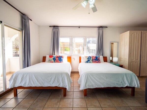 Habitación de hotel con dos camas, ventilador de techo, ventana con cortinas, armario de madera y piso de baldosas.