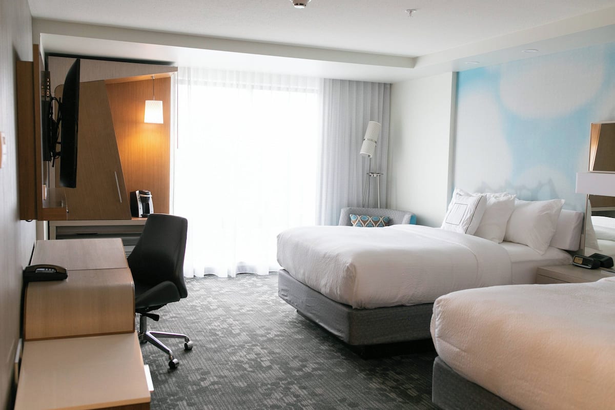 A hotel room with a bed, a desk, a chair, a lamp, and a large window.