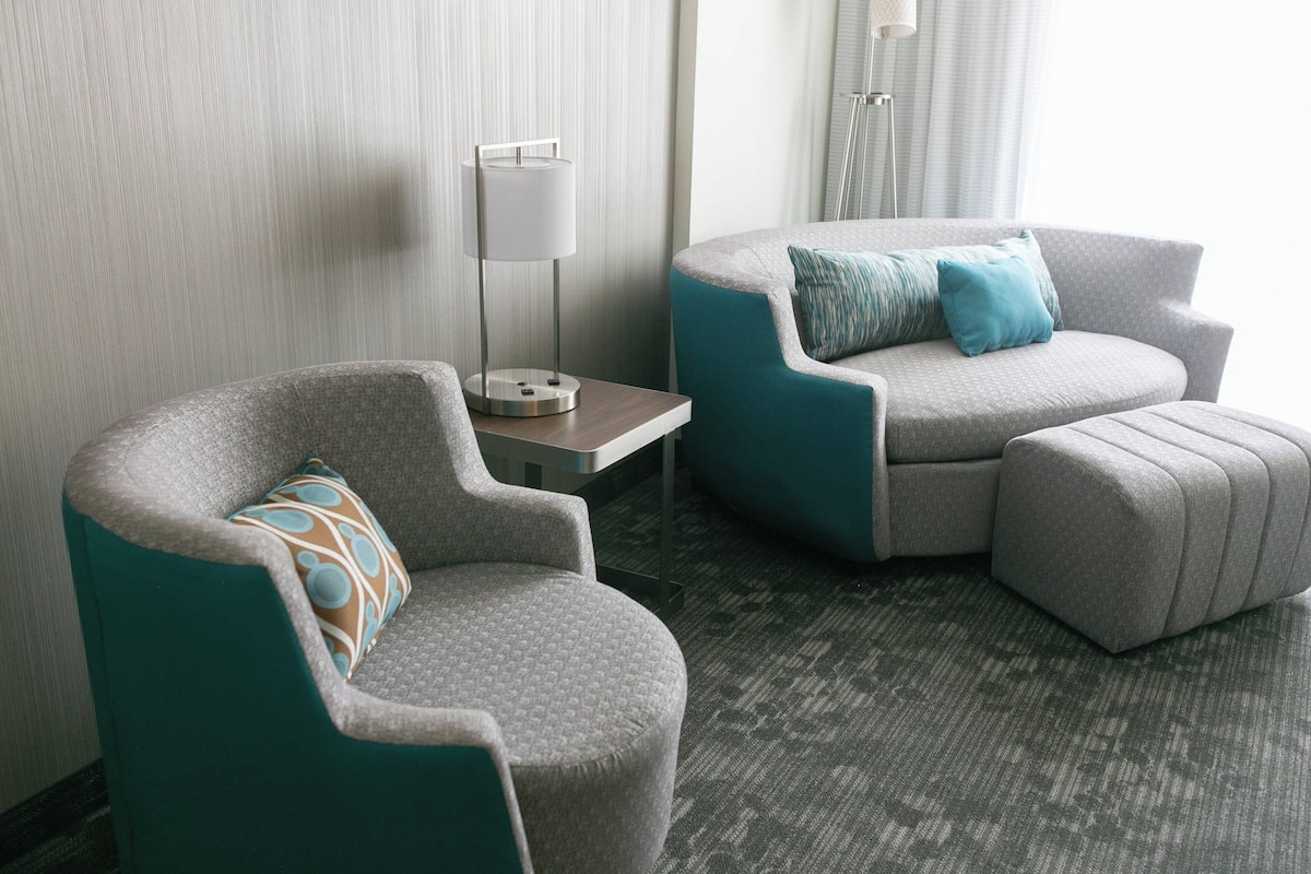 A modern hotel room with a grey armchair, a teal armchair, and a matching ottoman. A floor lamp is on a side table.