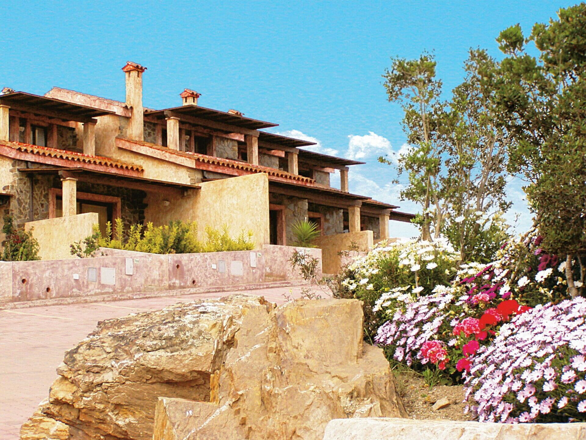 Un edificio grande y rústico con varios niveles y fachada de piedra, rodeado de un jardín paisajístico con plantas en flor y una roca grande al frente.