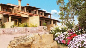 Un edificio grande e rustico con diversi livelli e una facciata in pietra, circondato da un giardino curato con piante fiorite e un grosso masso in primo piano.
