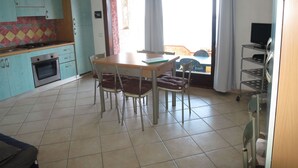 Una cucina con mobili color petrolio, un tavolo da pranzo con quattro sedie, una TV su un mobile basso e vista attraverso una porta scorrevole in vetro.