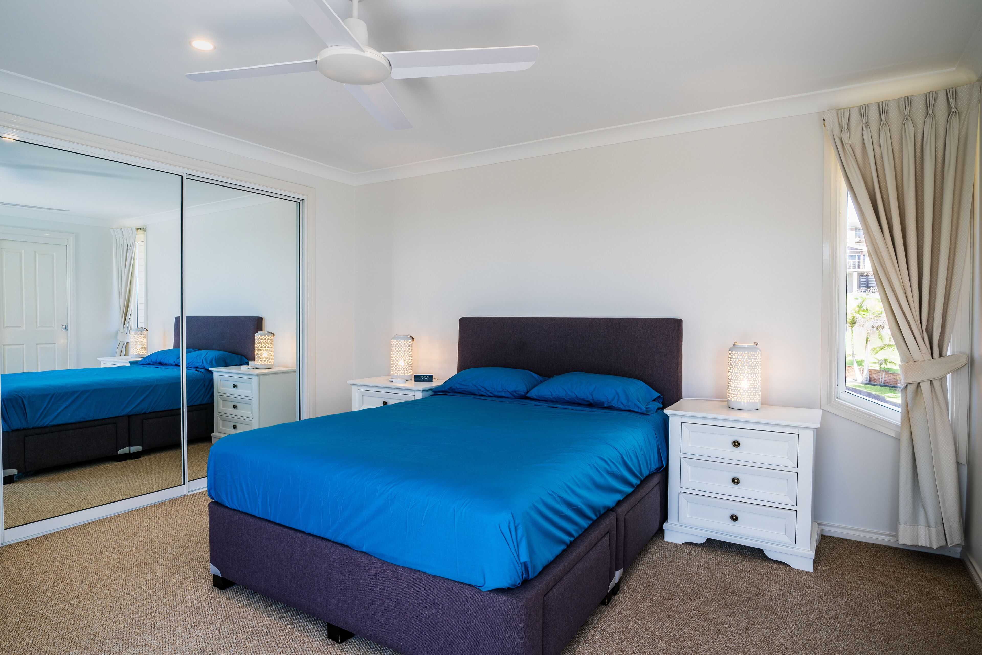 A bedroom with a large bed, a nightstand with a lamp, a ceiling fan, a window with curtains, and a mirror.