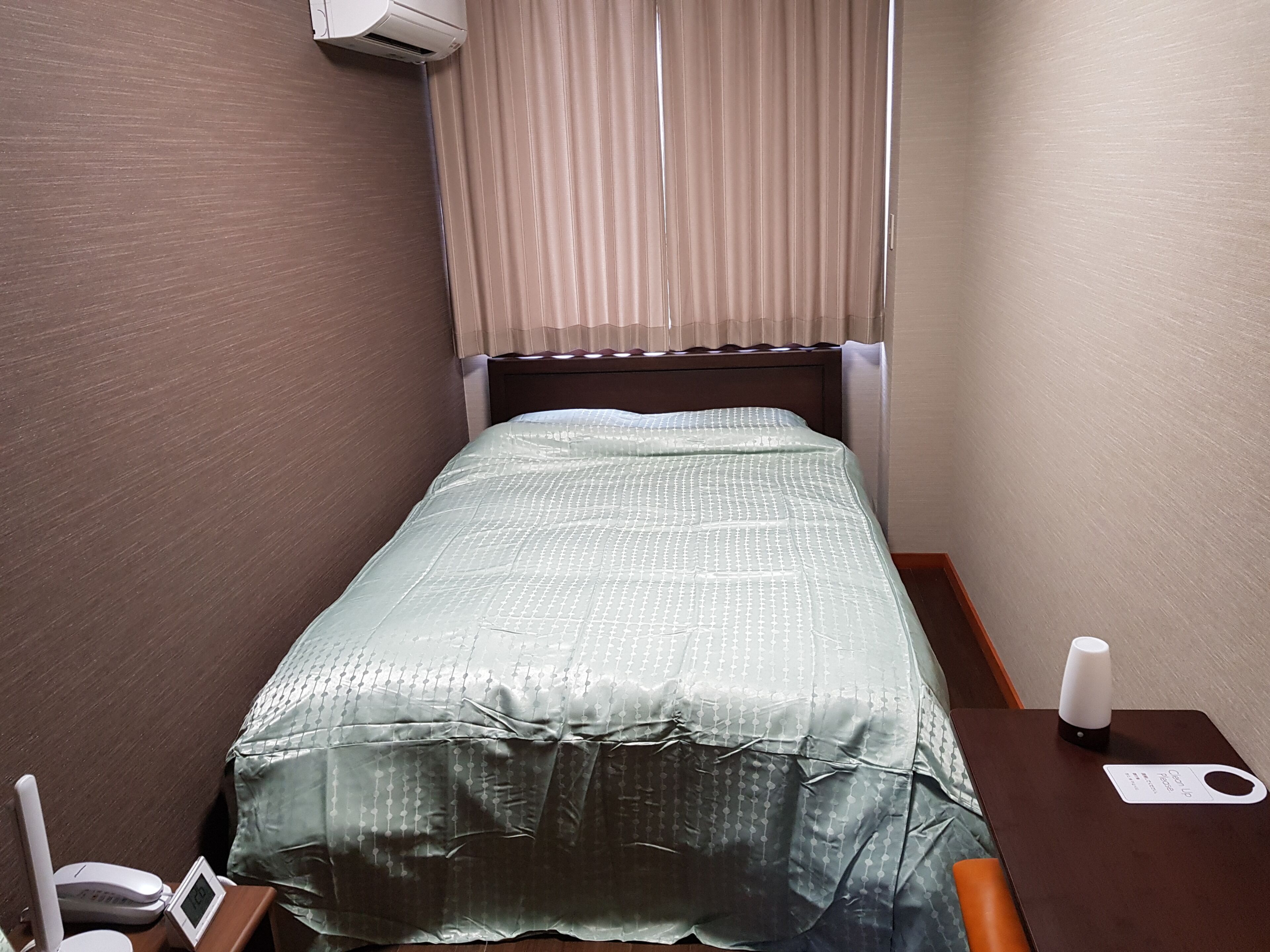 A small, neatly made bed with a patterned bedspread, a bedside table with a lamp, and a wall-mounted air conditioner.
