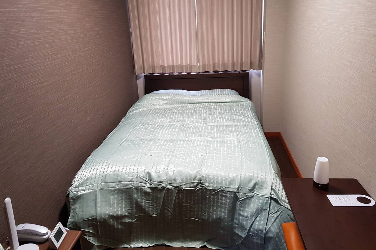 Uma cama pequena e bem arrumada, com um cobre-leito estampado, uma mesinha de cabeceira com abajur e um ar-condicionado fixado na parede.