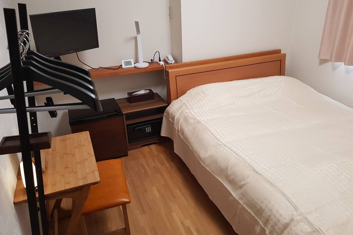 Quarto de hotel com cama, escrivaninha e cadeira, televisão, mesa de cabeceira com telefone e luminária, e um cofre pequeno.