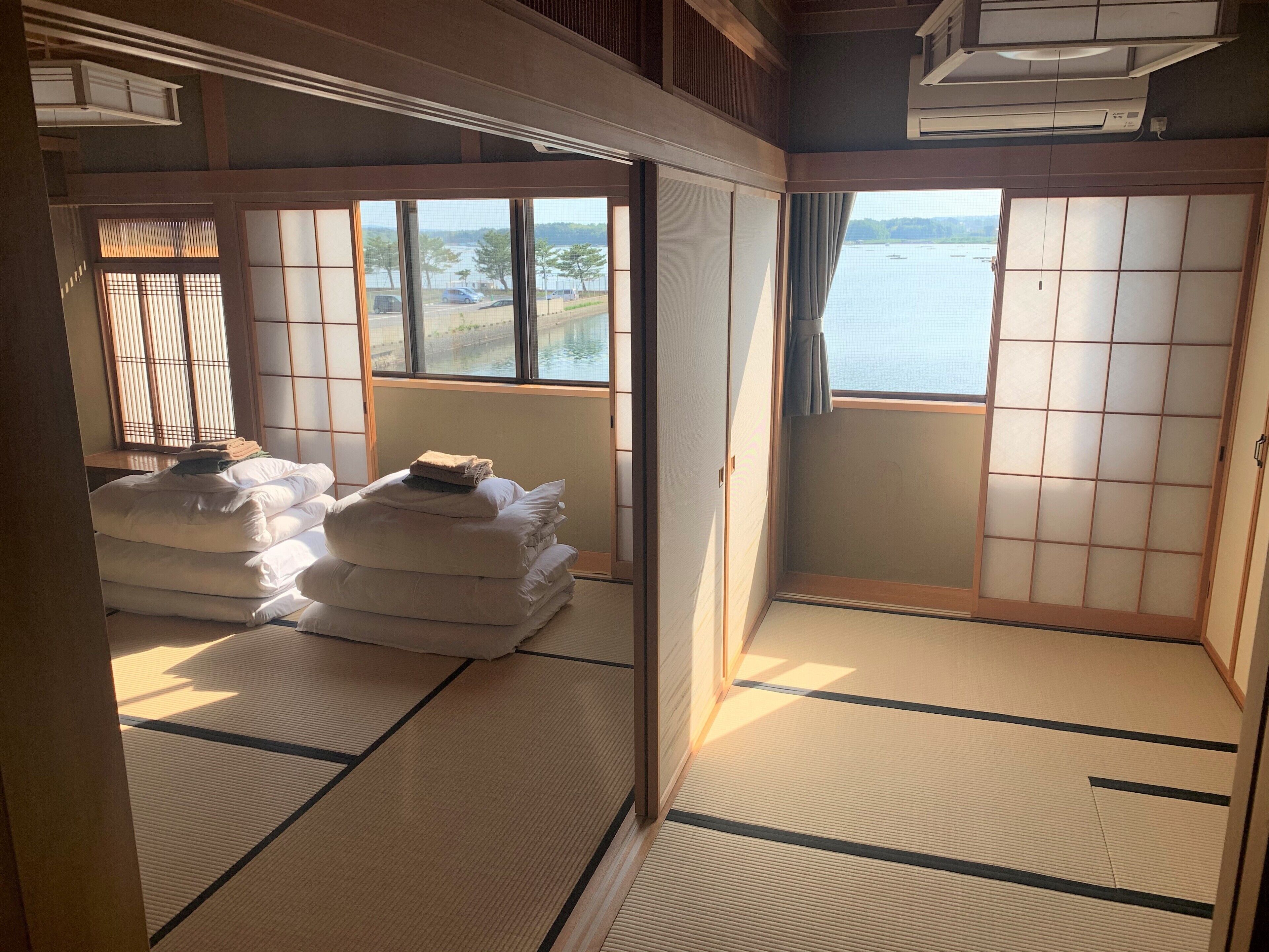 A traditional Japanese room with tatami flooring, a bed with white bedding, a view of a body of water through large windows, and sliding doors.