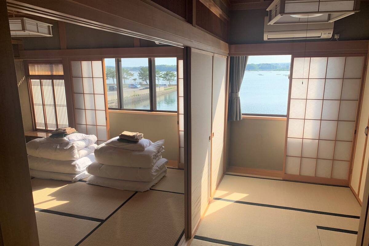 Um quarto japonês tradicional com tatames, uma cama com roupa de cama branca, vista para um corpo de água através de grandes janelas e portas deslizantes.