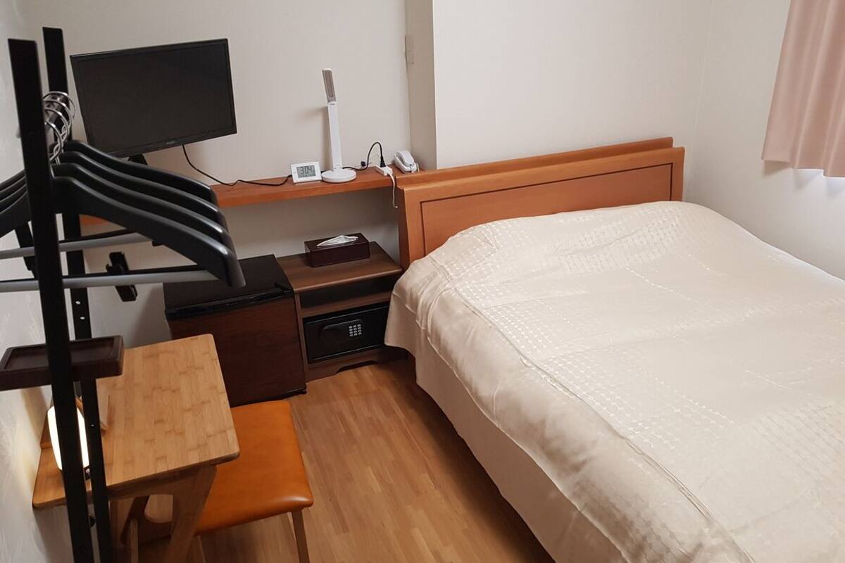 Quarto de hotel com cama, escrivaninha com cadeira, televisão, abajur e telefone num criado-mudo.