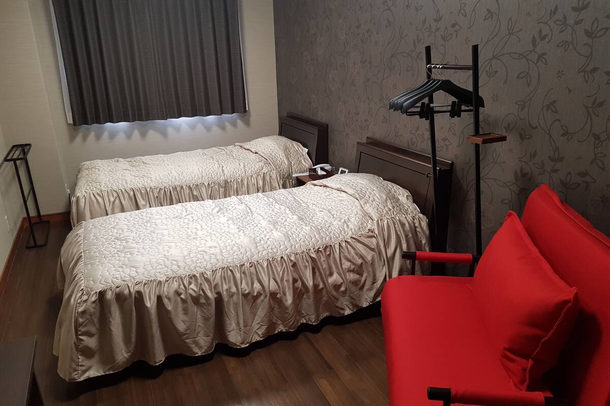 Quarto com duas camas, uma cadeira vermelha, um abajur e uma janela com cortinas.