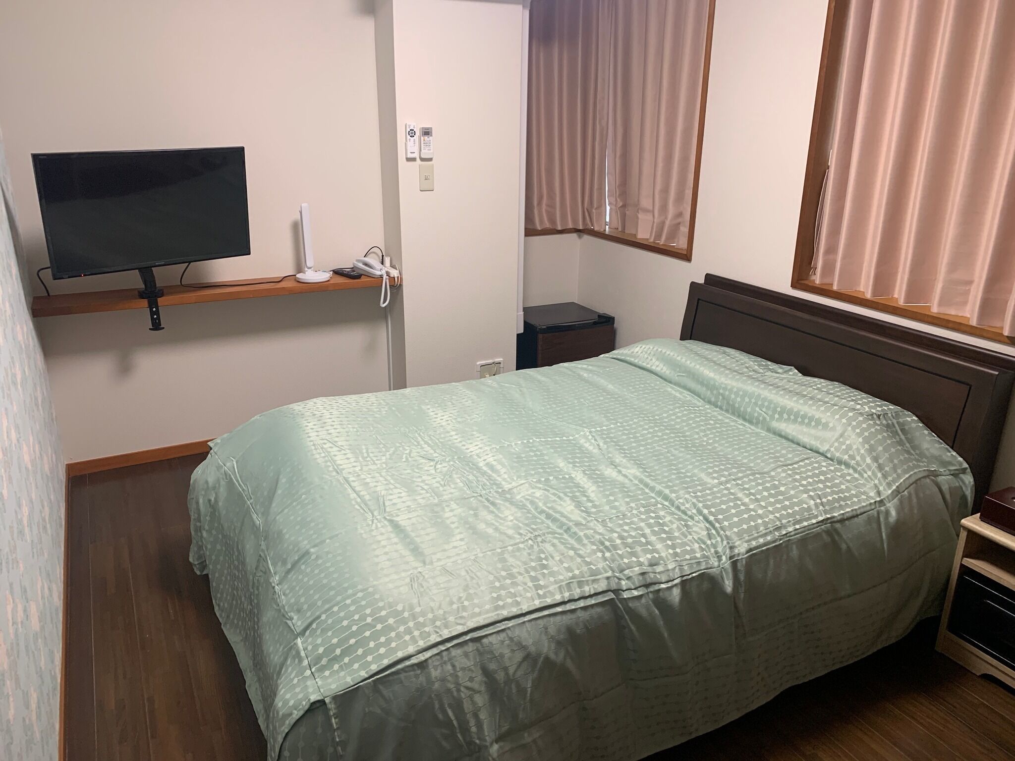 A bedroom with a bed, a TV on a shelf, a bedside table, a window with curtains, and a wall-mounted thermostat.