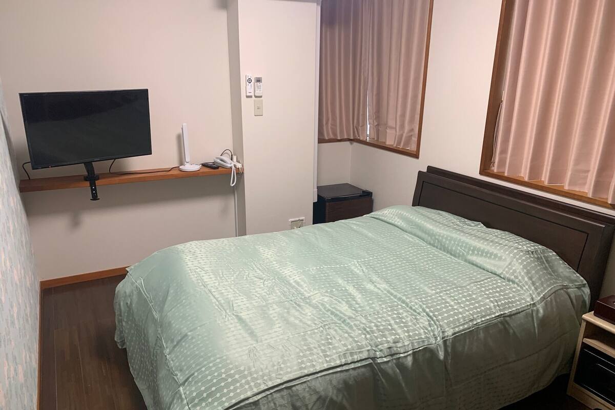 Quarto com cama, televisão em prateleira, mesa de cabeceira, janela com cortinas e termostato fixado na parede.