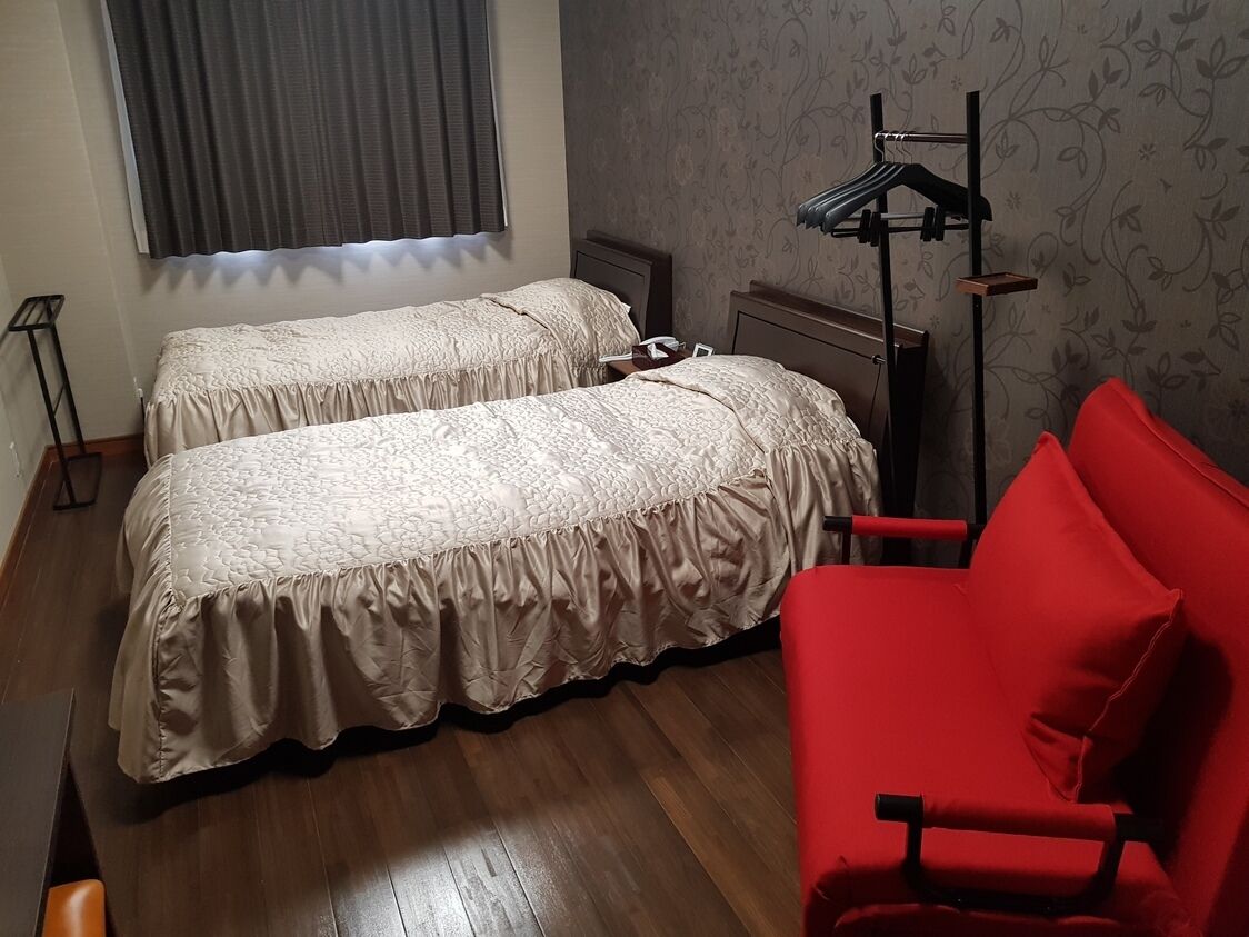A room with two beds, a red chair, a standing lamp, and a radiator.