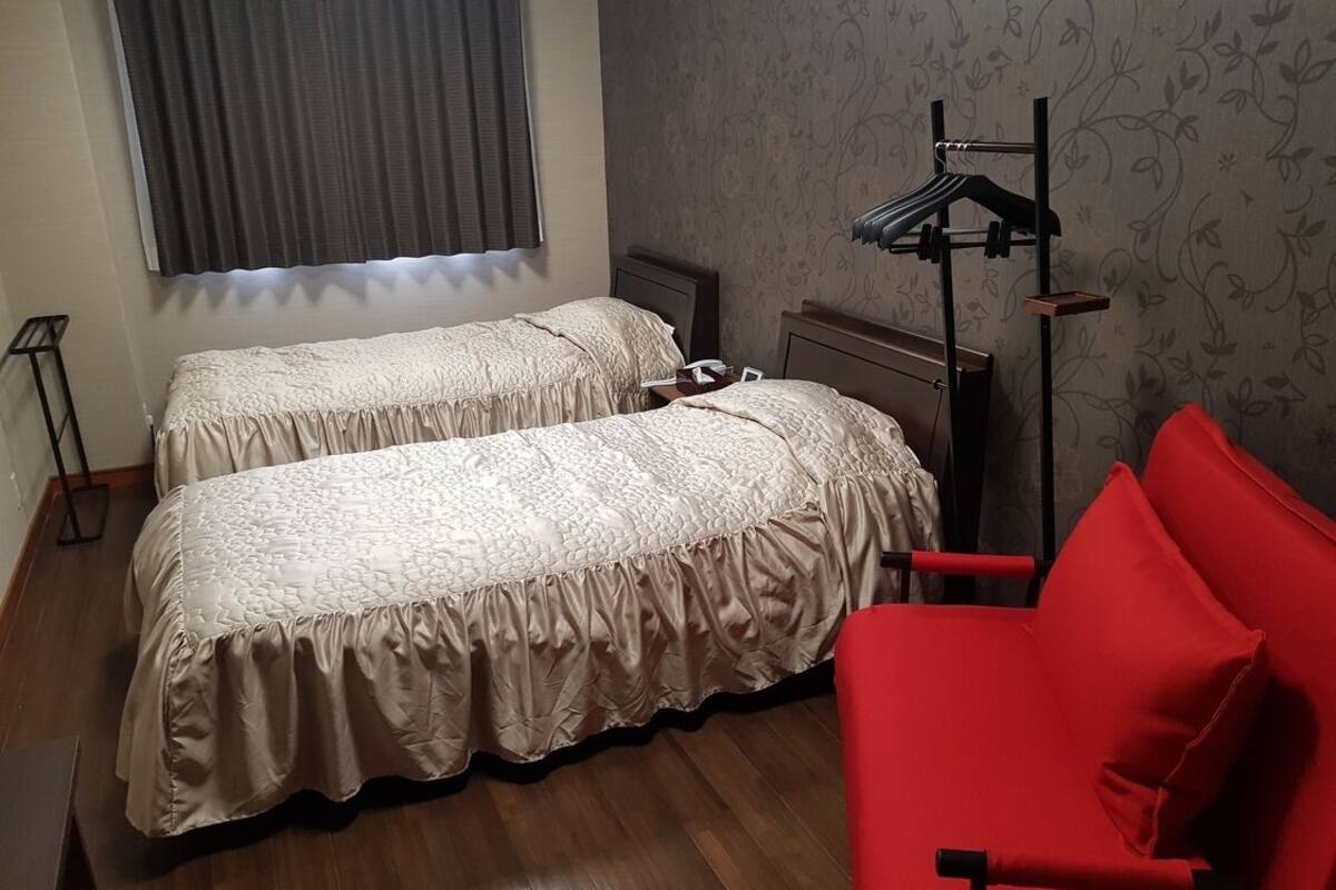 Quarto com duas camas, uma cadeira vermelha, um candeeiro e um radiador.