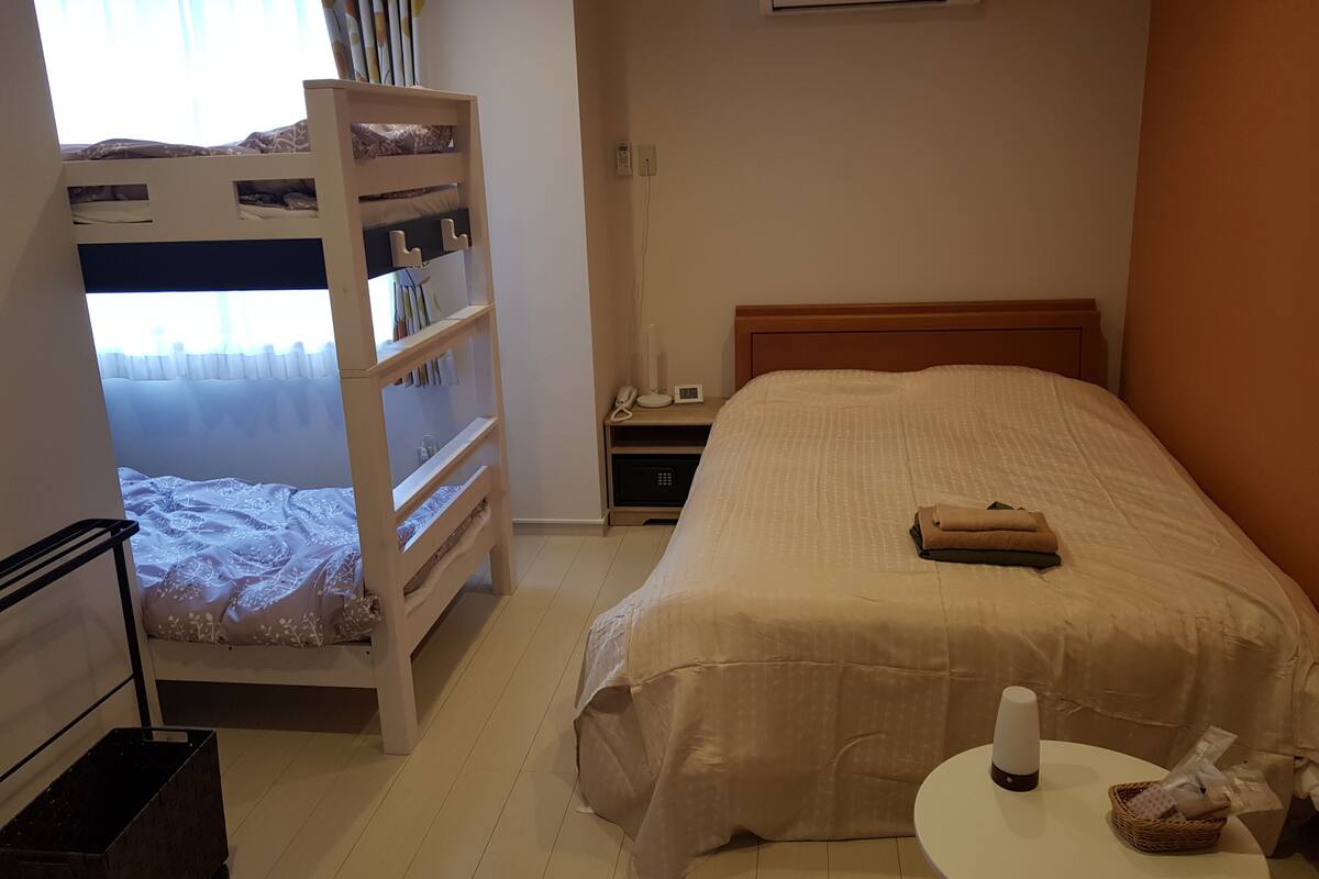 Um quarto com cama beliche, uma cama de solteiro, mesa de cabeceira com abajur, telefone e uma pequena mesa com uma garrafa e uma cesta.