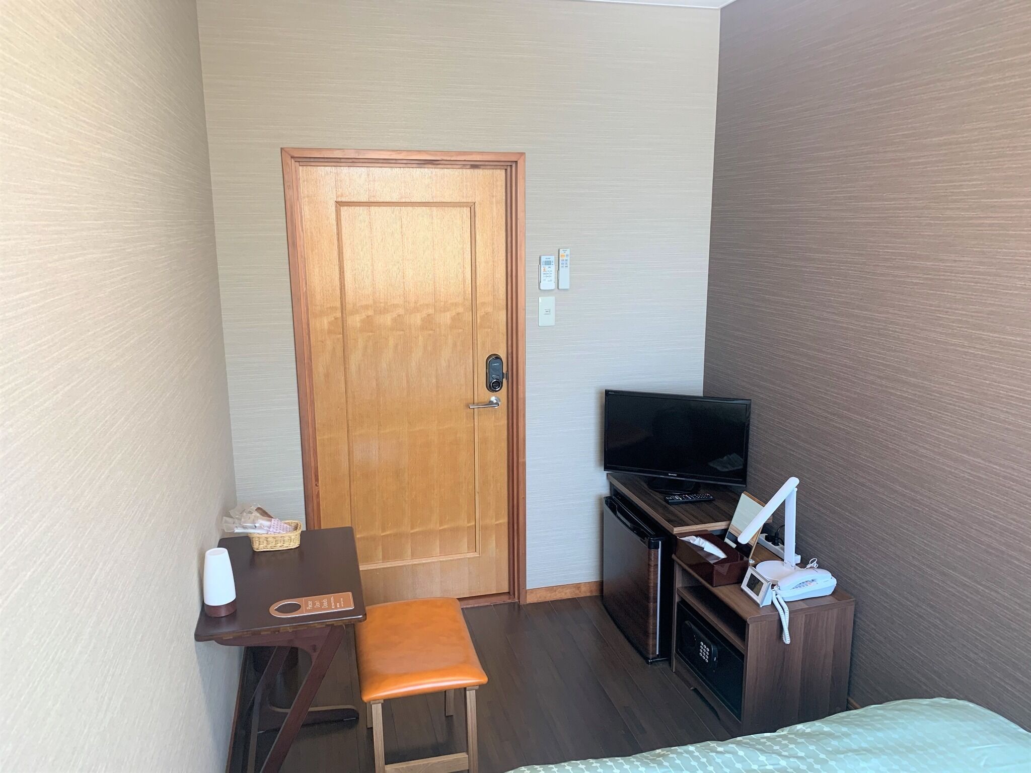 A hotel room with a wooden door, a desk with a chair, a television, a minibar, and a bed.