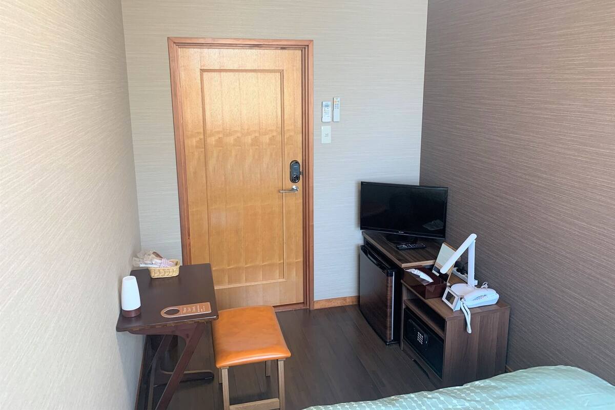 Quarto de hotel com porta de madeira, escrivaninha com cadeira, televisão, minibar e uma cama.