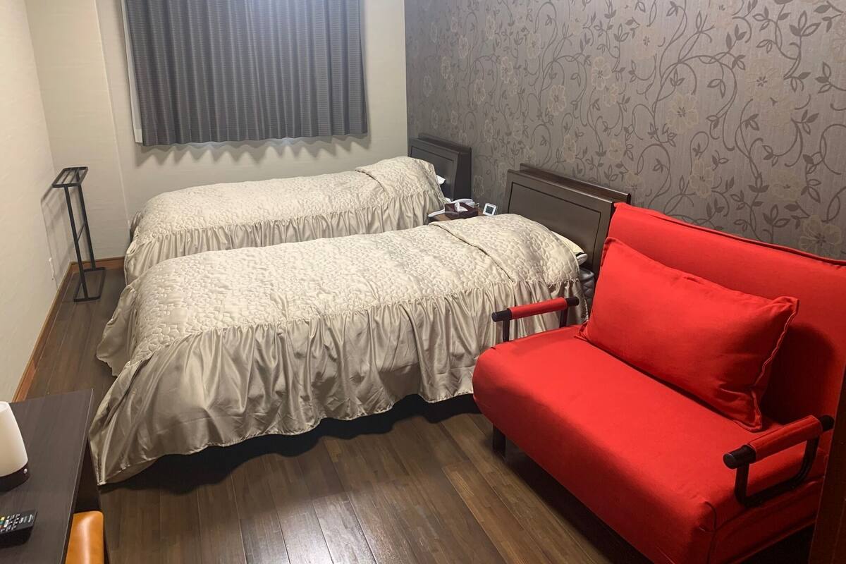Quarto com duas camas, um sofá vermelho, uma mesa pequena e uma janela com cortinas.