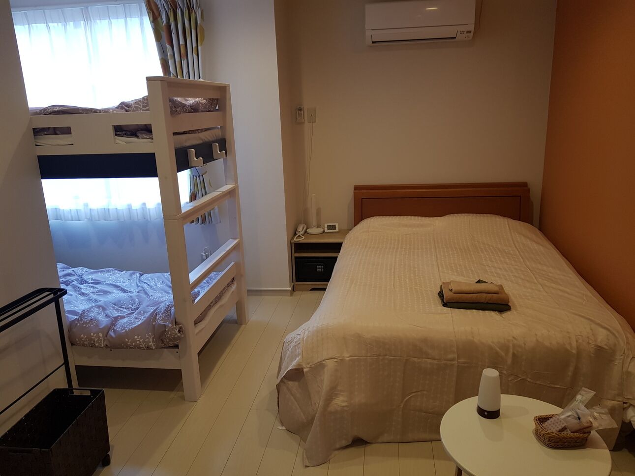 A hotel room with a bunk bed, a single bed, a bedside table with a lamp, a small table with a basket, and an air conditioning unit on the wall.