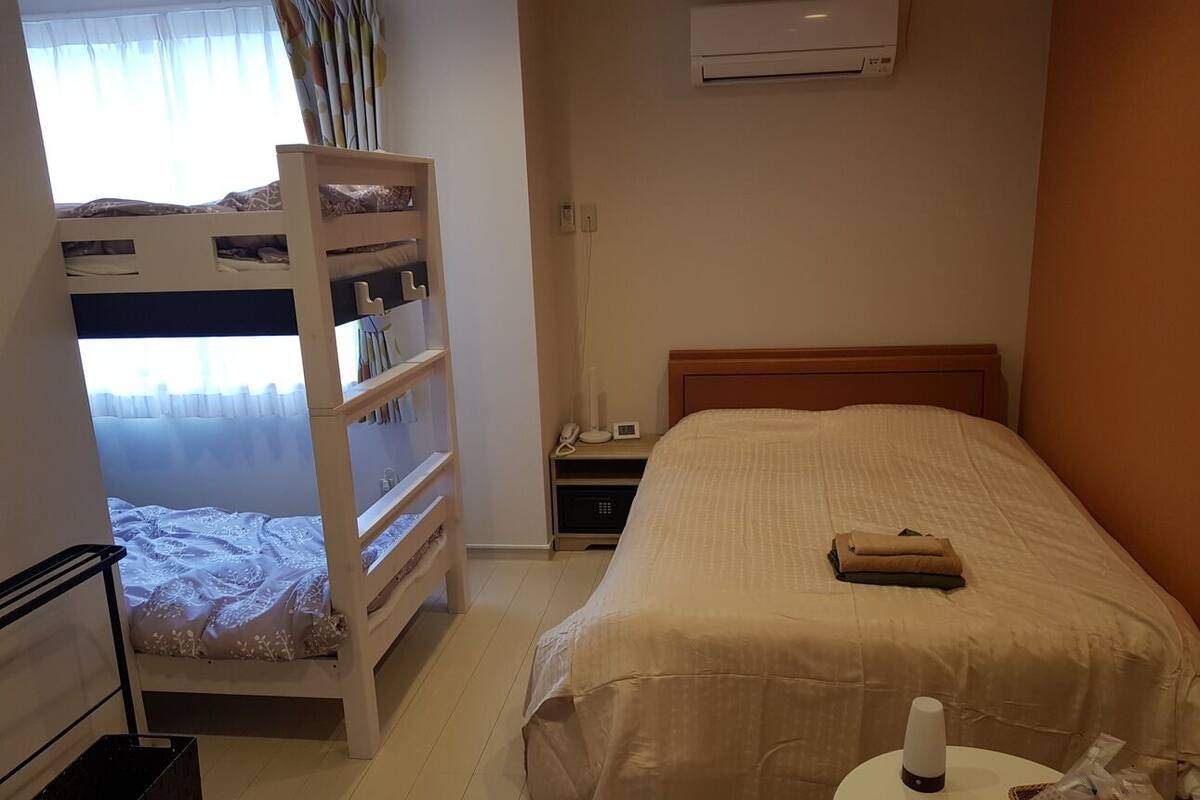 Quarto de hotel com cama beliche, uma cama de solteiro, mesa de cabeceira com abajur, pequena mesa com cesta e ar condicionado na parede.